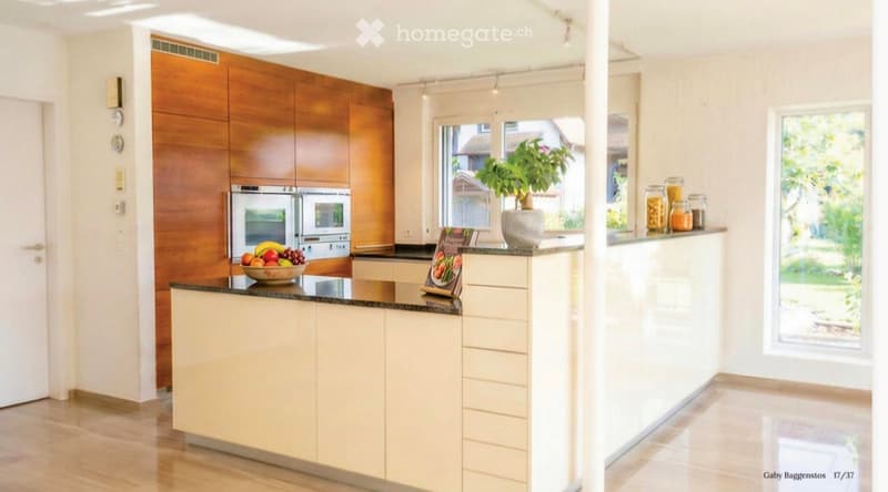 Modern kitchen with light-colored cabinets, a central island, and a black countertop. Features include an oven, a fruit bowl, a potted plant, and several jars on the counter.