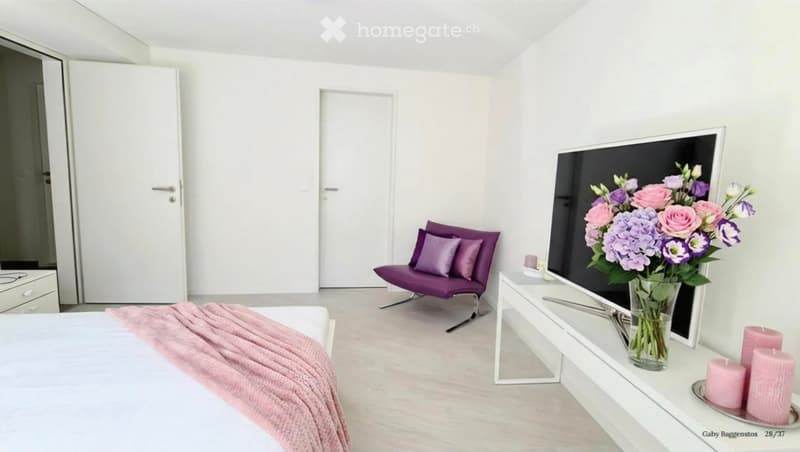 Modern bedroom with a white bed, pink throw, purple chair, white side table, vase with flowers, and candles.