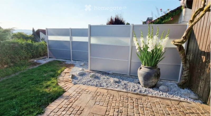 Garden area with a modern grey fence, a large ceramic vase with white lilies, a paved brick pathway, and a small grassy patch.