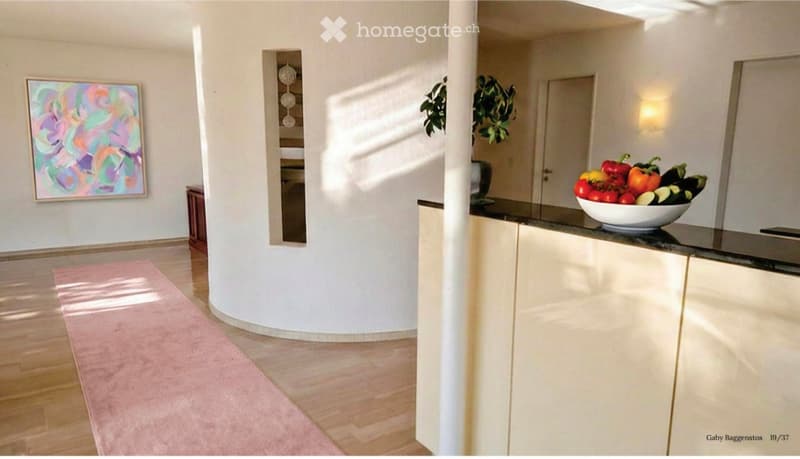 Modern open-plan area with a kitchen counter, a bowl of vegetables, a painting on the wall, and a plant in the corner.