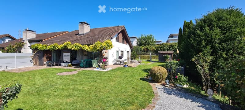 Two-story house with a tiled roof, white exterior, and well-maintained garden featuring a lawn, patio seating, and various plants.