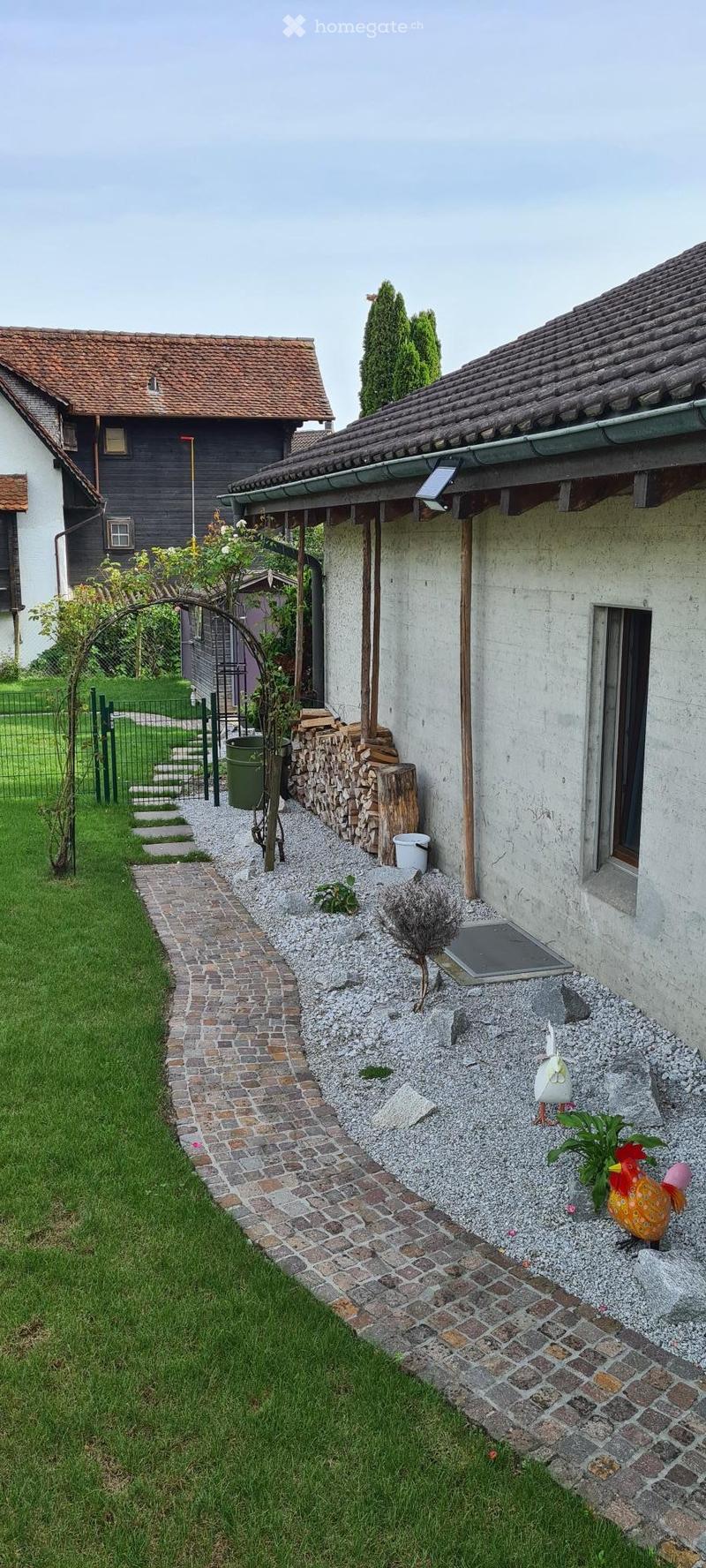 Garden area with a brick pathway leading to a house, featuring a stack of firewood, a small decorative rooster, and a green trash bin.