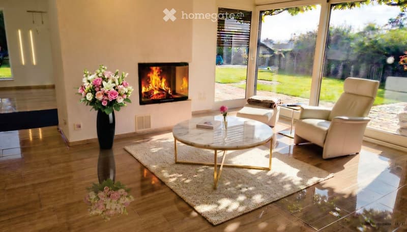 Modern living room with a fireplace, large windows offering a view of the garden, a white leather sofa, a round marble coffee table, and decorative vases with flowers.