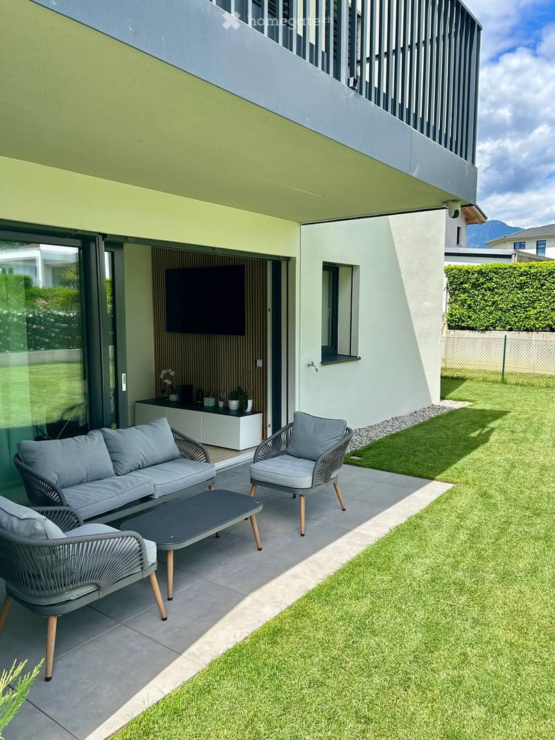Modern outdoor seating area with a grey sofa, two matching chairs, and a coffee table, adjacent to a well-maintained lawn and modern building.