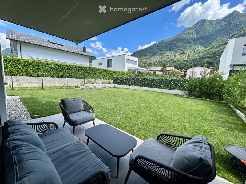 A well-maintained garden with a neatly trimmed lawn, surrounded by a low stone wall and a hedge. The patio area features a grey sofa, two wicker chairs, and a small black coffee table, all set against a backdrop of a scenic mountain view.