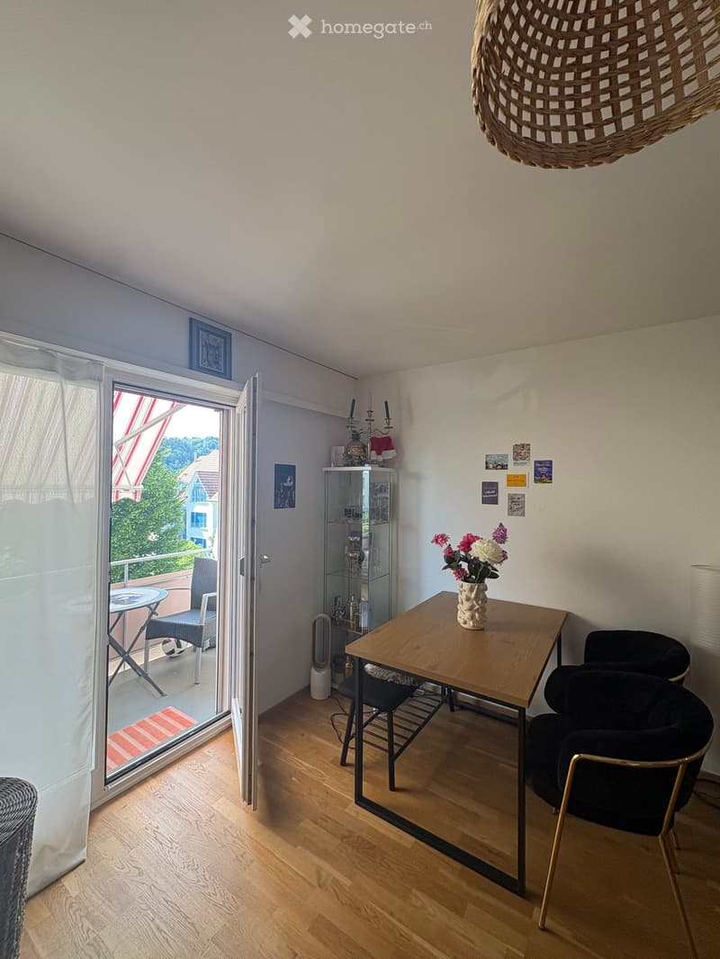 Bright living room with wooden floor, large window leading to a balcony, a wooden table with chairs, a vase with flowers, a candelabra, and a glass cabinet.