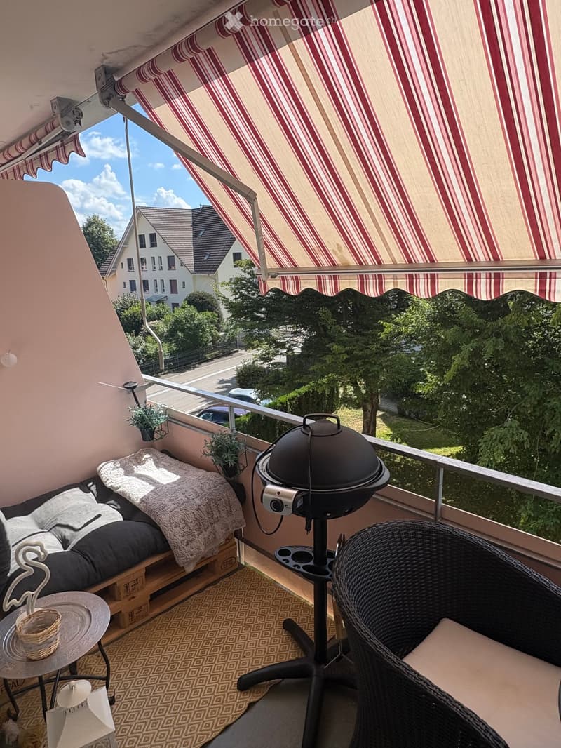 Balcony with a striped awning, a wicker chair, a small table, a grill, and a cushioned seating area.