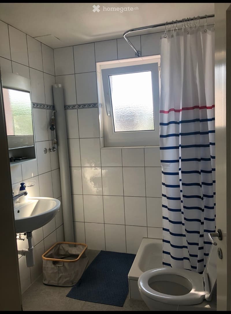 Bathroom with a sink, toilet, bathtub, and shower curtain. Features tiled walls, a window, and a small trash bin.
