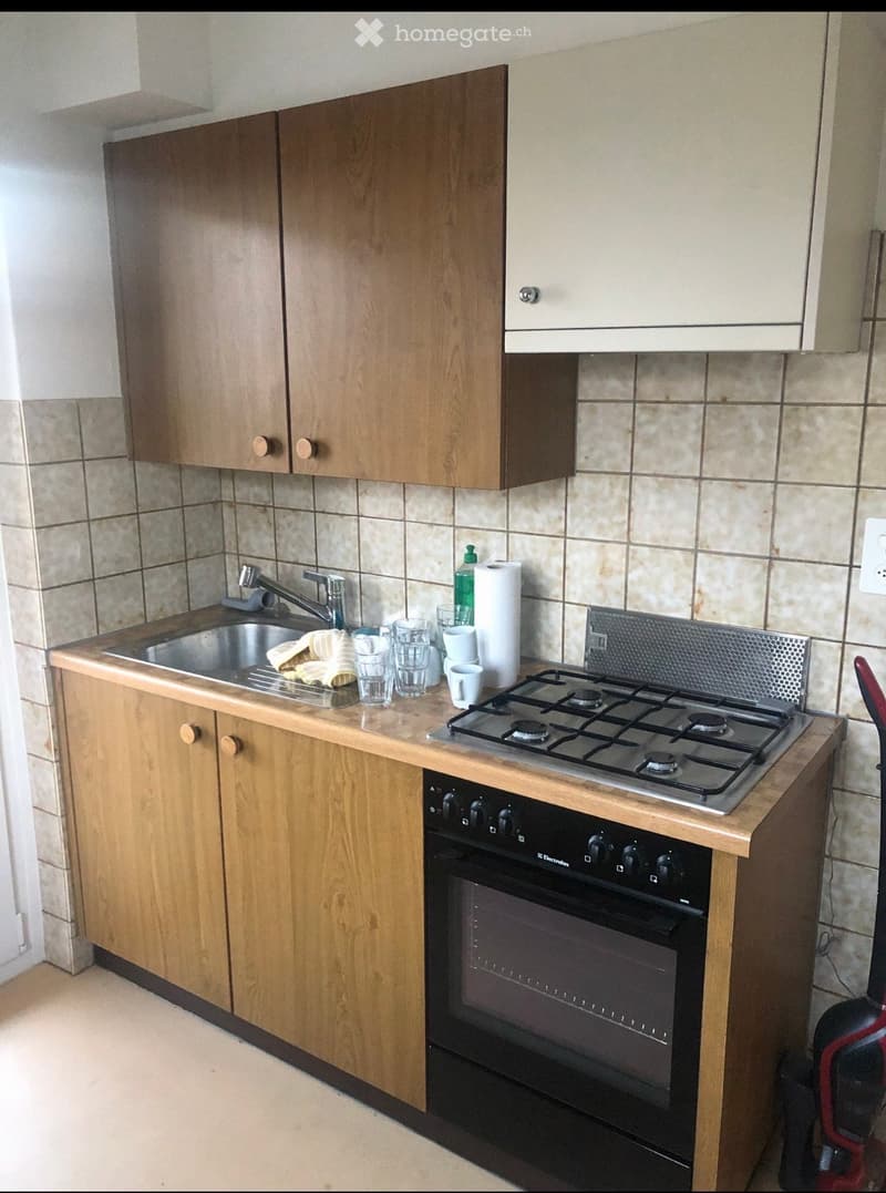 A modern kitchen with wooden cabinets, a stainless steel sink, a gas stove, and an oven. The countertop holds several cups and a bottle of dish soap. The walls are tiled with beige tiles.