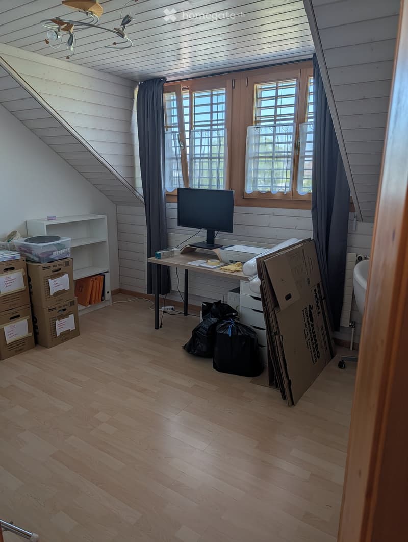 A room with wooden flooring, a slanted ceiling, and large windows. It contains a table with a screen, printer, and chair, along with shelves and boxes.