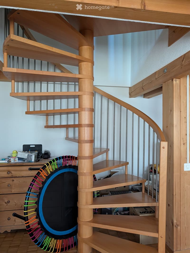Spiral staircase with wooden steps and railings, adjacent to a wooden dresser with drawers and a colorful exercise wheel on the floor.