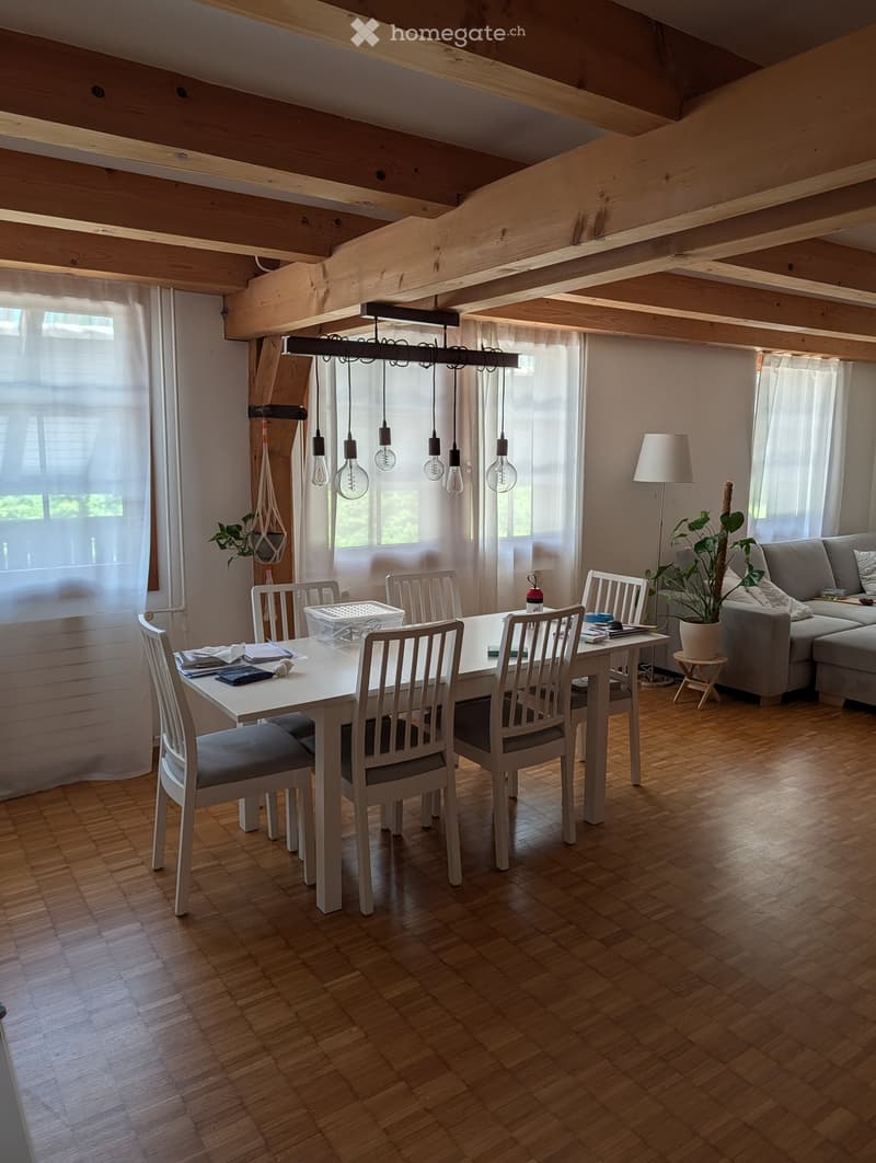 Dining area with a white dining table, six chairs, a grey sofa, floor lamp, and potted plants. Wooden ceiling beams and large windows with white curtains.