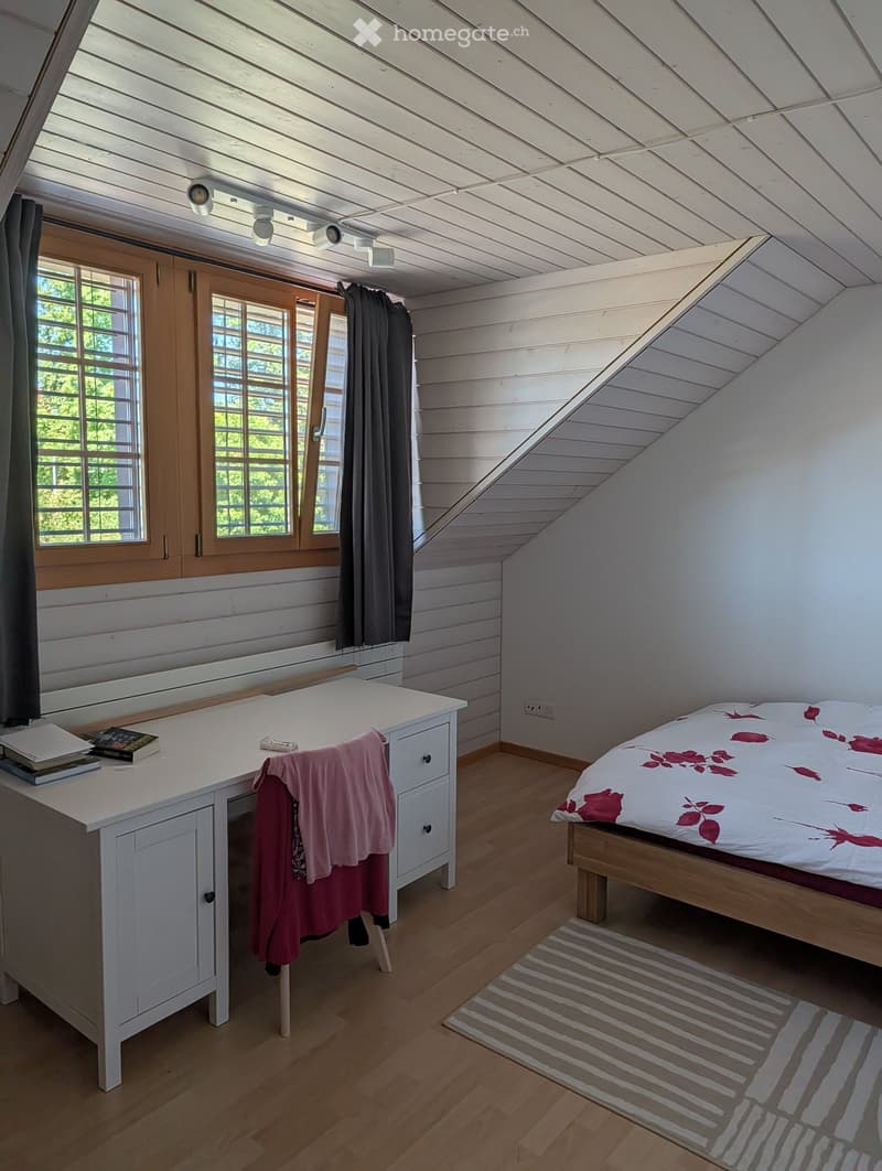 Cozy attic bedroom with wooden slanted ceilings, large windows, a bed with a floral duvet, a white desk with books and a pink jacket, and a striped rug on the floor.