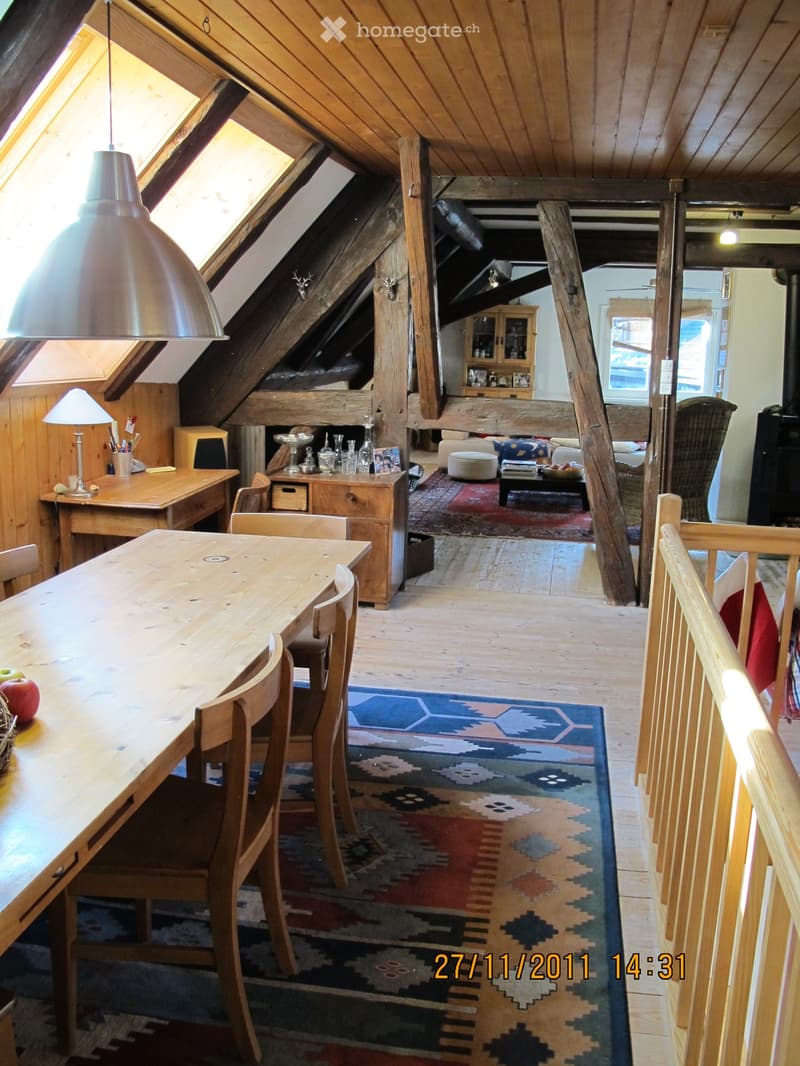 A spacious attic room with wooden beams and exposed rafters, featuring a long wooden dining table with chairs, a colorful patterned rug, a desk with a lamp, and a living area with a sofa, coffee table, and ottoman.