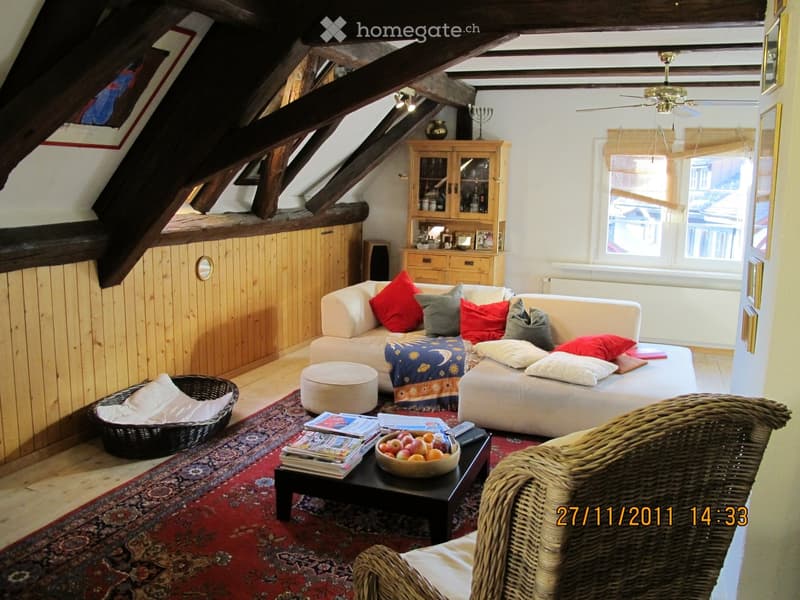 Cozy living room with exposed wooden beams, white sofa with colorful cushions, coffee table with fruit bowl and magazines, wicker chair, and a wicker basket on the floor.