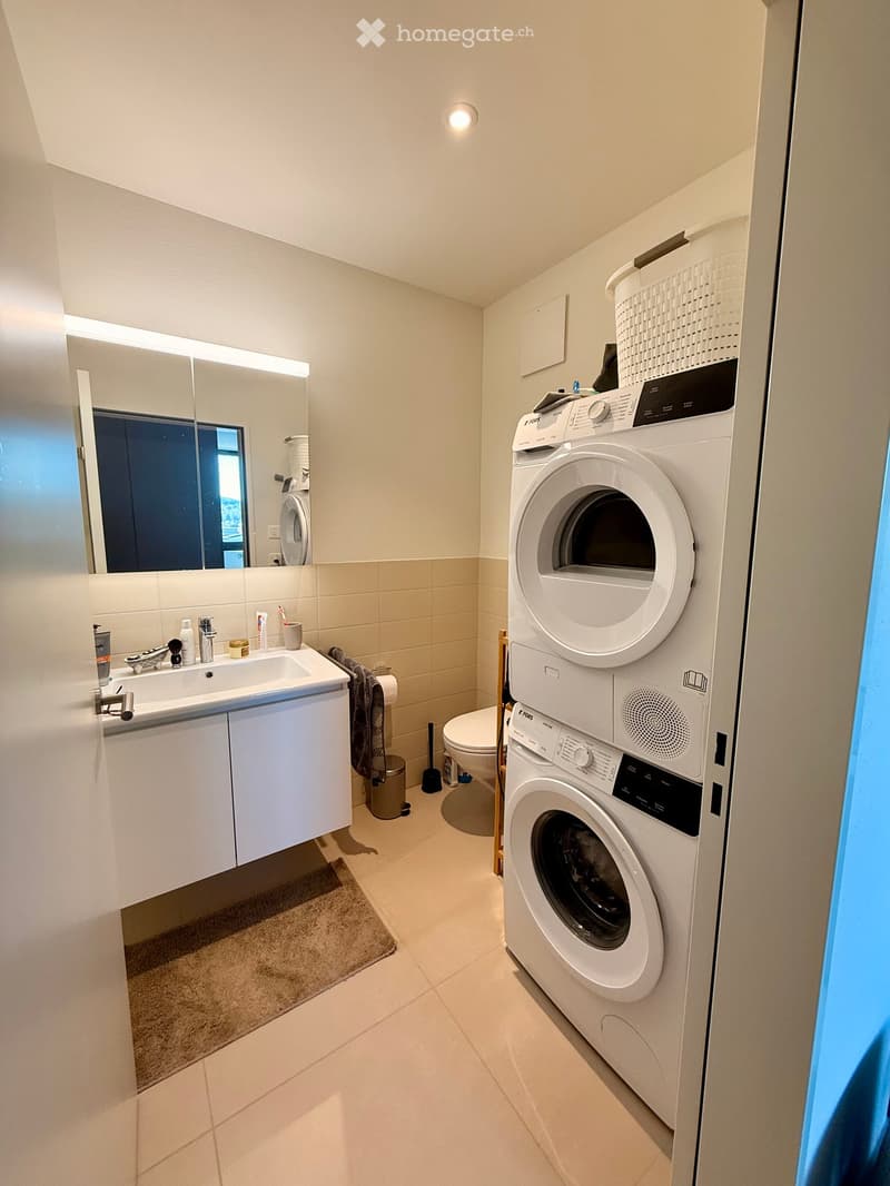 Modern bathroom with a sink, mirror, toilet, and stacked washer/dryer unit. Light-colored tiles and white cabinetry.