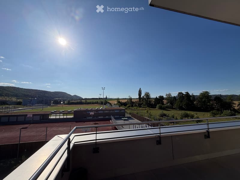 Balcony with metal railing overlooking a sports complex, including a running track, basketball court, and surrounding greenery.