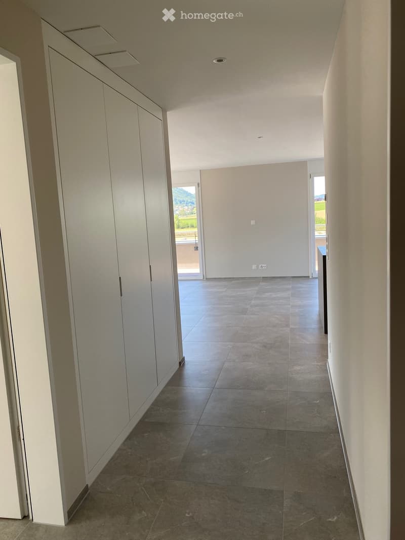 Empty hallway with tiled floor, white walls, and built-in storage units on one side. Natural light enters from a large window at the end.