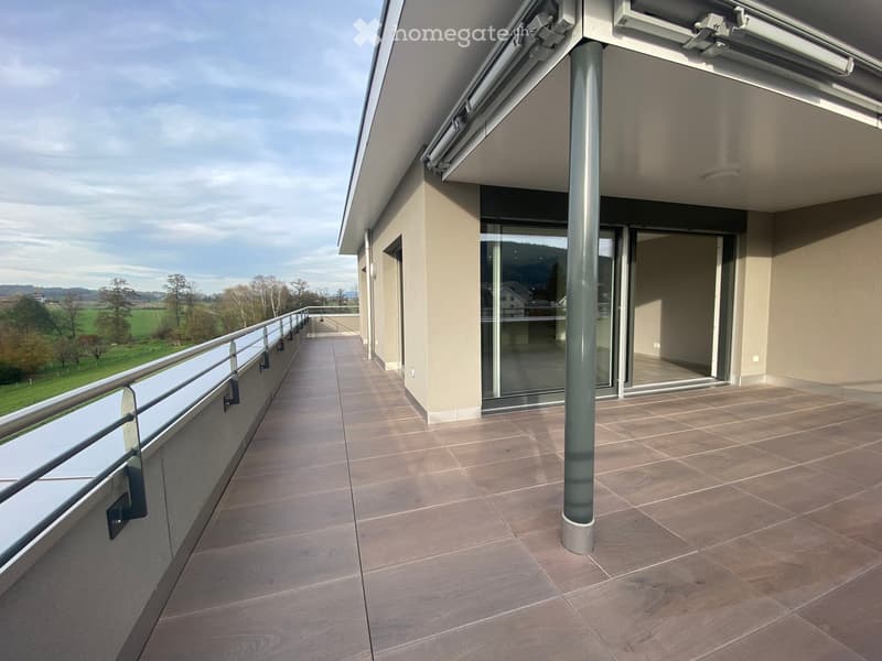 Balcony with tiled floor, glass railing, and open sliding doors leading to an indoor space. The view shows a green landscape with trees and fields.