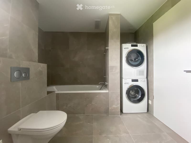 Modern bathroom with a toilet, bathtub, and stacked washing machine. Features include tiled walls and a sleek design.