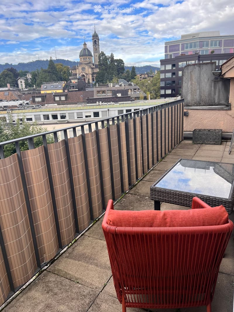 Balcony with a red wicker chair, a wicker table, and a wooden privacy screen, overlooking a cityscape with a church and surrounding buildings.
