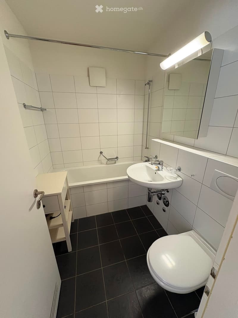 Modern bathroom with white tiles, a bathtub, a sink, a toilet, a mirror, and a small cabinet.