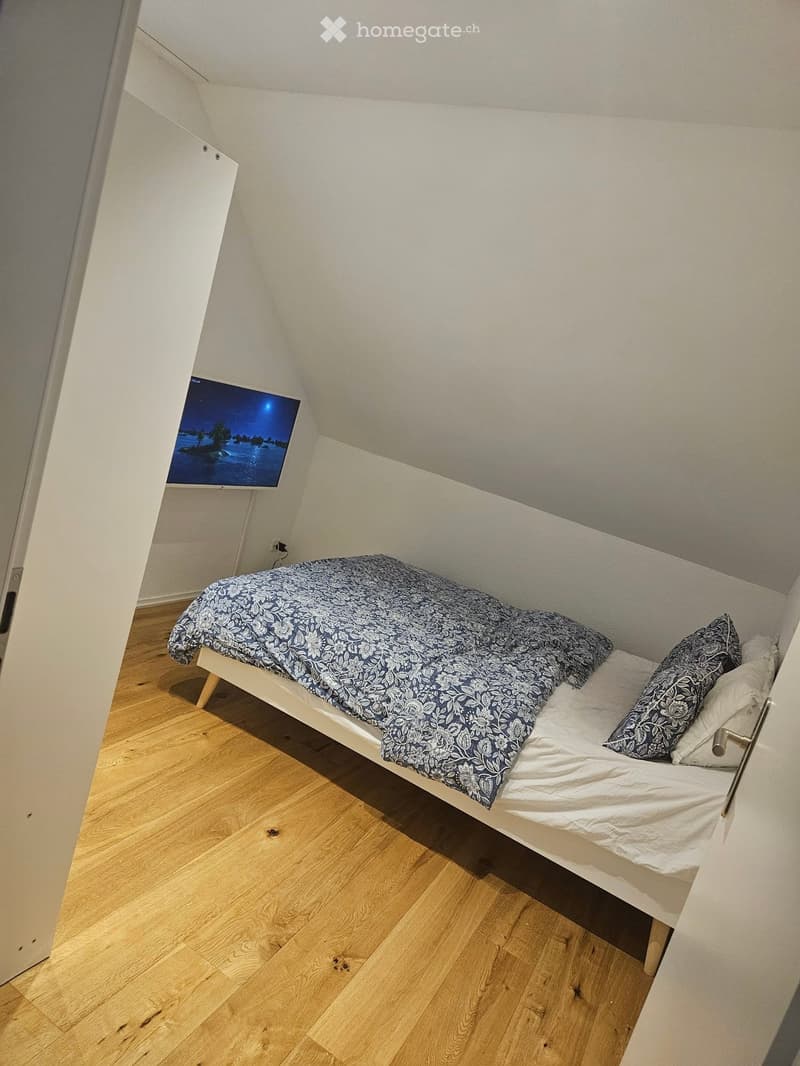 Cozy attic bedroom with wooden flooring, a single bed with patterned bedding, pillows, and a mounted television displaying a scenic image.