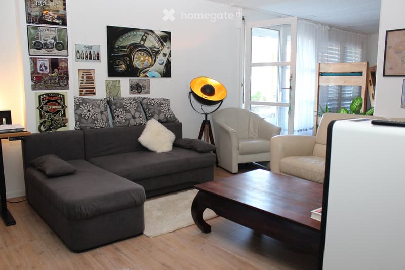 Living room with a dark grey sectional sofa, two white armchairs, a wooden coffee table, a floor lamp, and several posters on the wall.