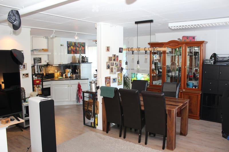 A modern, open-plan living and dining area with a wooden dining table and chairs, a kitchen area with white cabinets, a sink, and various appliances, and a wooden cabinet with glass doors.