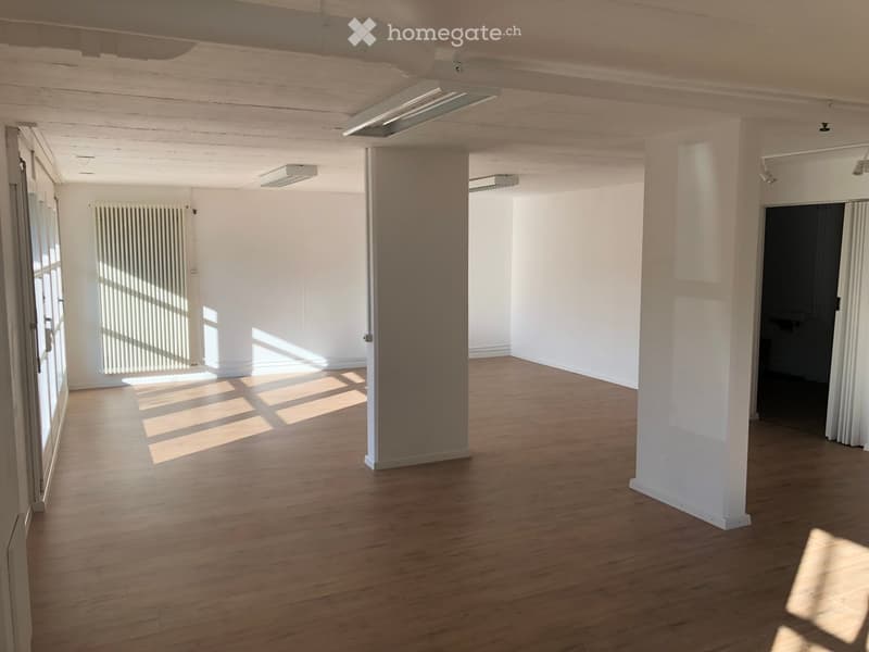 Empty room with wooden flooring, white walls, and large windows allowing natural light. Features include a radiator, a door, and exposed ceiling beams.