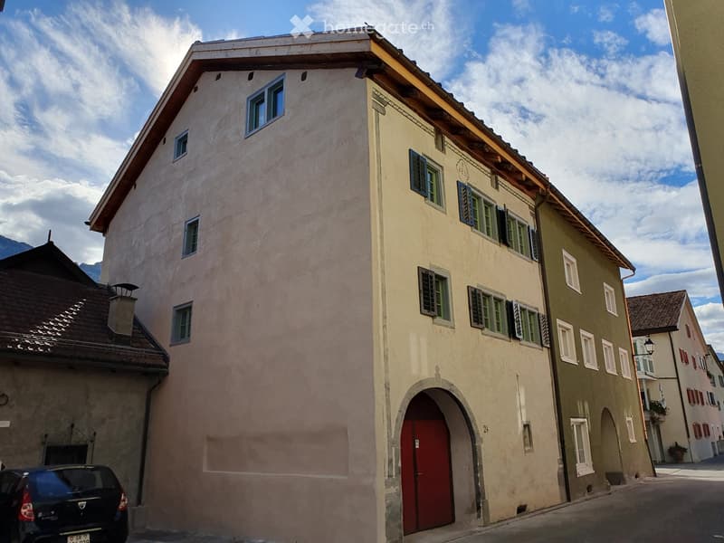 A multi-story building with beige walls, green shutters, and a red door, situated in a narrow street with neighboring buildings and a parked car.