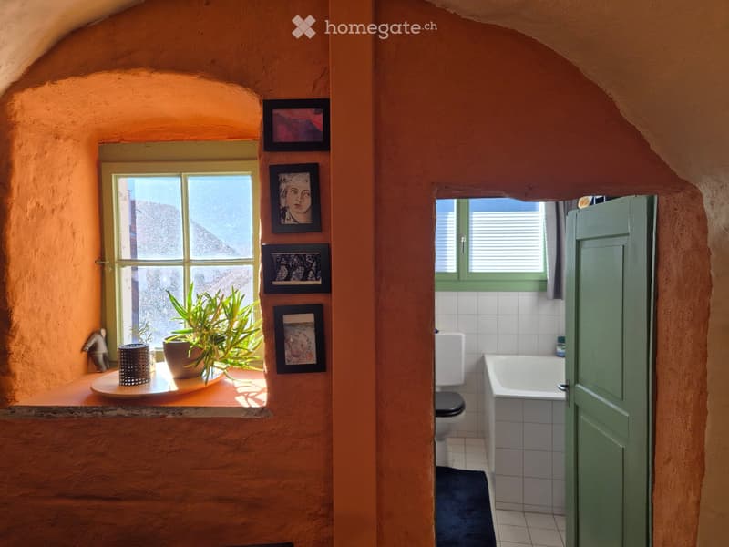 Bathroom with a toilet, bathtub, and a small windowed sitting area with a plant and framed pictures on the wall.