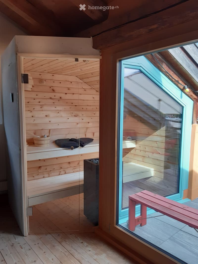 Wooden sauna room with a glass door leading to a wooden deck, featuring a red bench, a wooden bucket, and towels.