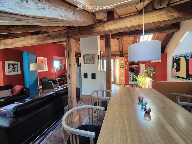 Open-plan living and dining area with exposed wooden beams, a black leather sofa, wooden dining table, chairs, floor lamp, and artwork on the walls.