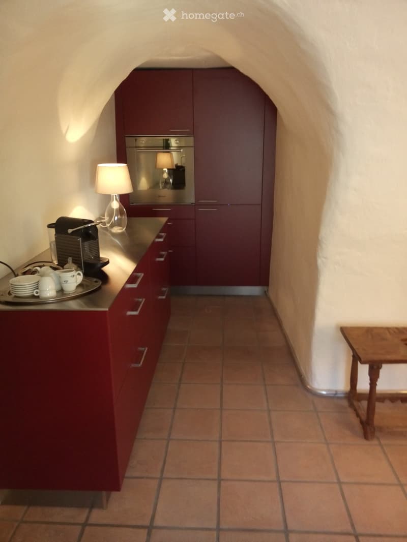 Modern kitchen with dark red cabinets, stainless steel oven, coffee maker, and a small wooden stool near the doorway.