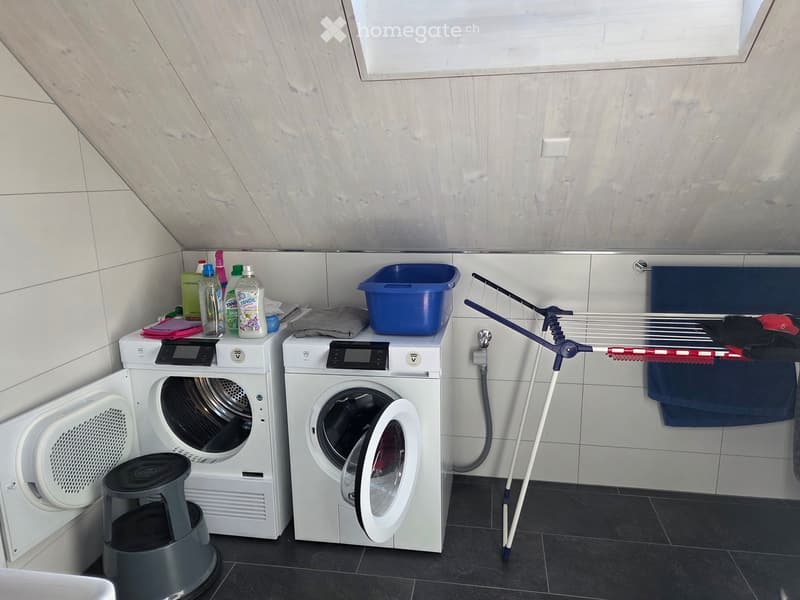 Laundry area with a washing machine, dryer, laundry basket, drying rack, and cleaning supplies on a shelf.