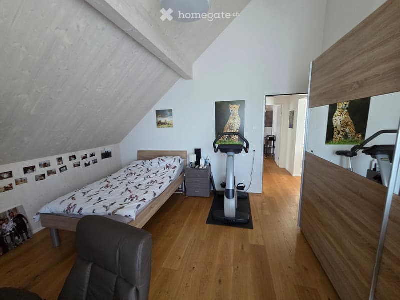 Cozy attic bedroom with wooden flooring, slanted ceiling, bed, nightstand, exercise bike, and a large mirror reflecting part of the room.