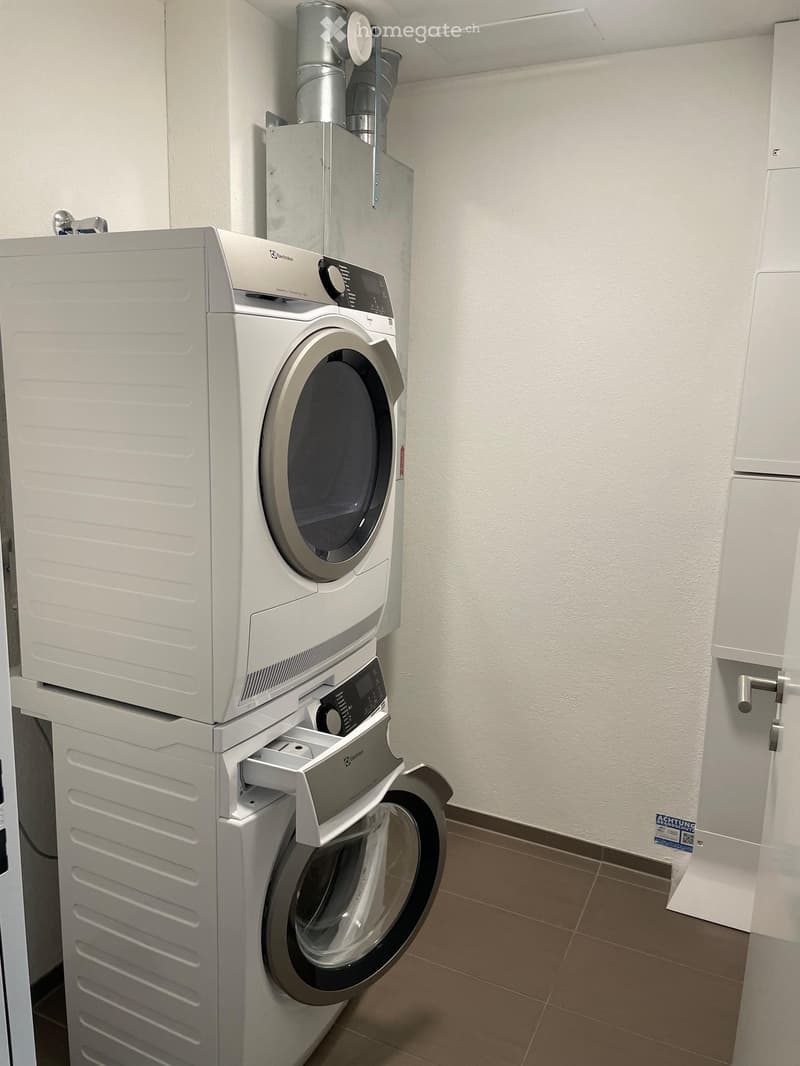 A small room with a white washing machine and dryer stacked vertically, white walls, and brown tiled floor.