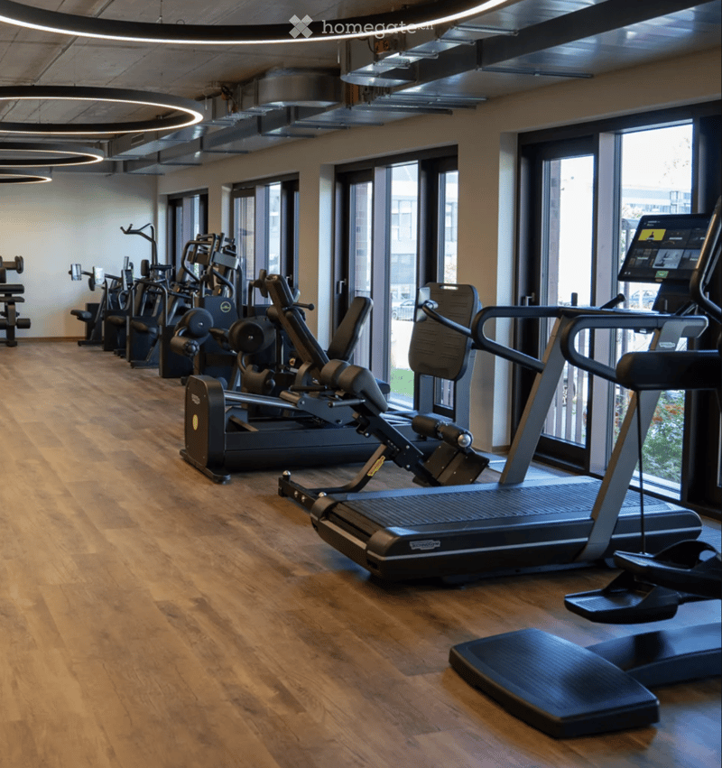 Modern fitness room with wooden flooring, large windows, and various exercise equipment including treadmills, elliptical machines, and weight machines.