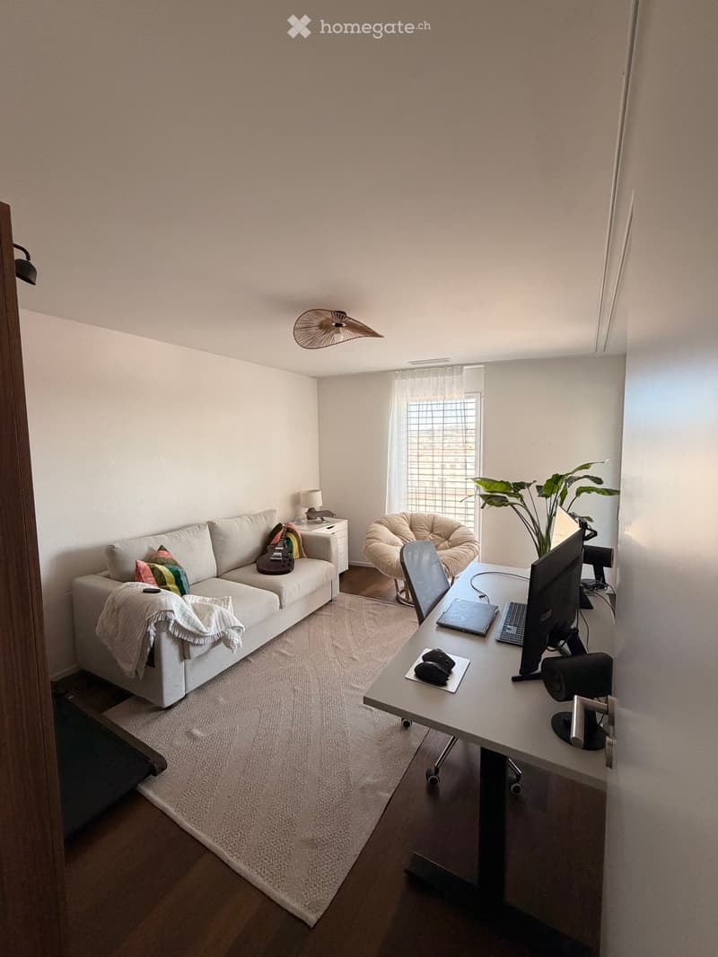 Modern living room with a white sofa, laptop desk, wicker chair, and guitar. Features a large window with white curtains and a plant in the corner.