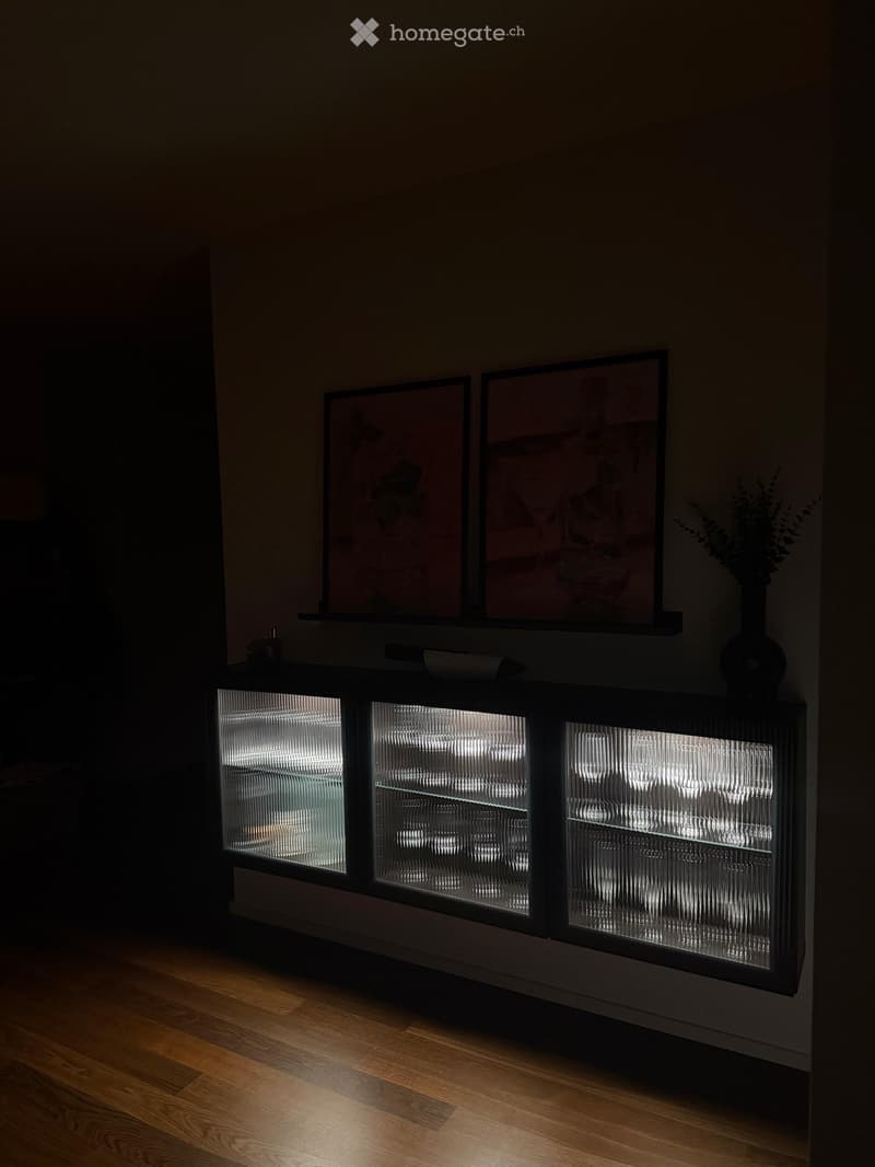 A modern, dimly lit room with a sleek black cabinet featuring glass doors, illuminated from within, displaying various items. Two framed pictures are mounted above the cabinet, and a tall vase with dried flowers sits on top.