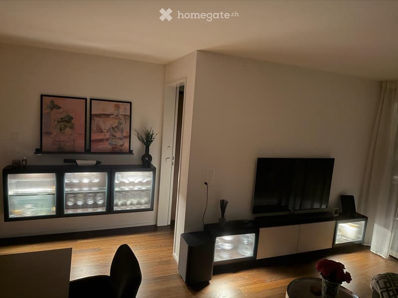 Modern living room with wooden flooring, a large flat-screen TV on a black stand, a sofa, a coffee table with a vase of flowers, and a cabinet with glass doors.