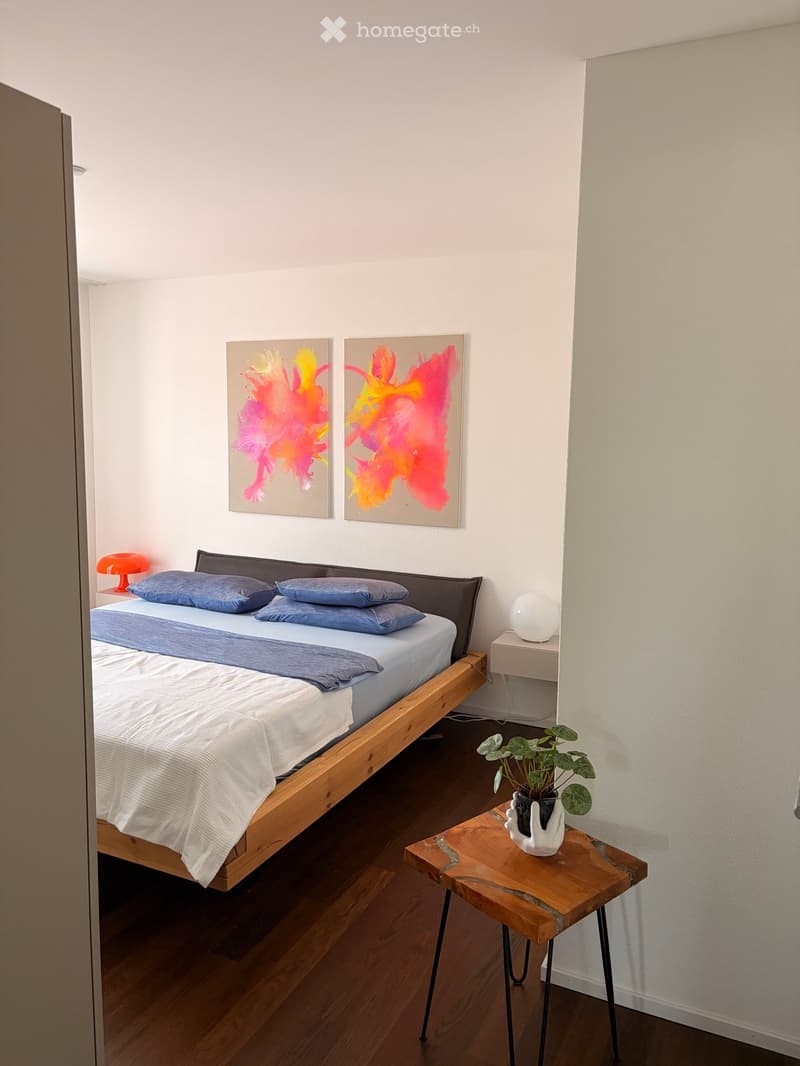 A modern bedroom with a wooden bed, blue bedding, nightstand with a lamp, small table with a plant, and colorful abstract artwork on the wall.
