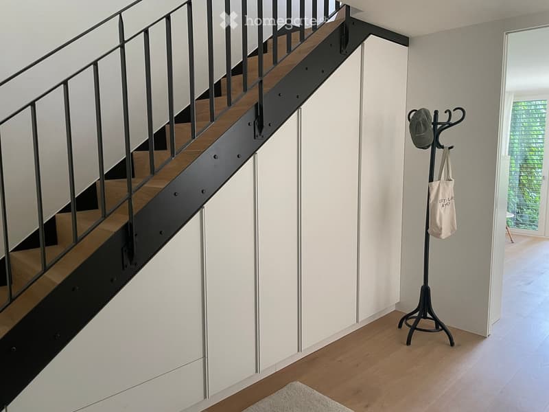 Modern hallway with wooden flooring, a staircase with wooden steps and black metal railing, a coat rack with a hat and bag, and a white rug on the floor.