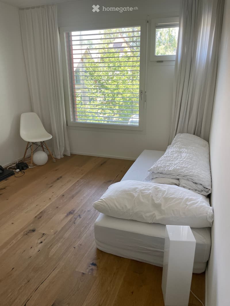 A modern bedroom with a wooden floor, a bed with white bedding, a white chair, and a small table near the window with curtains.