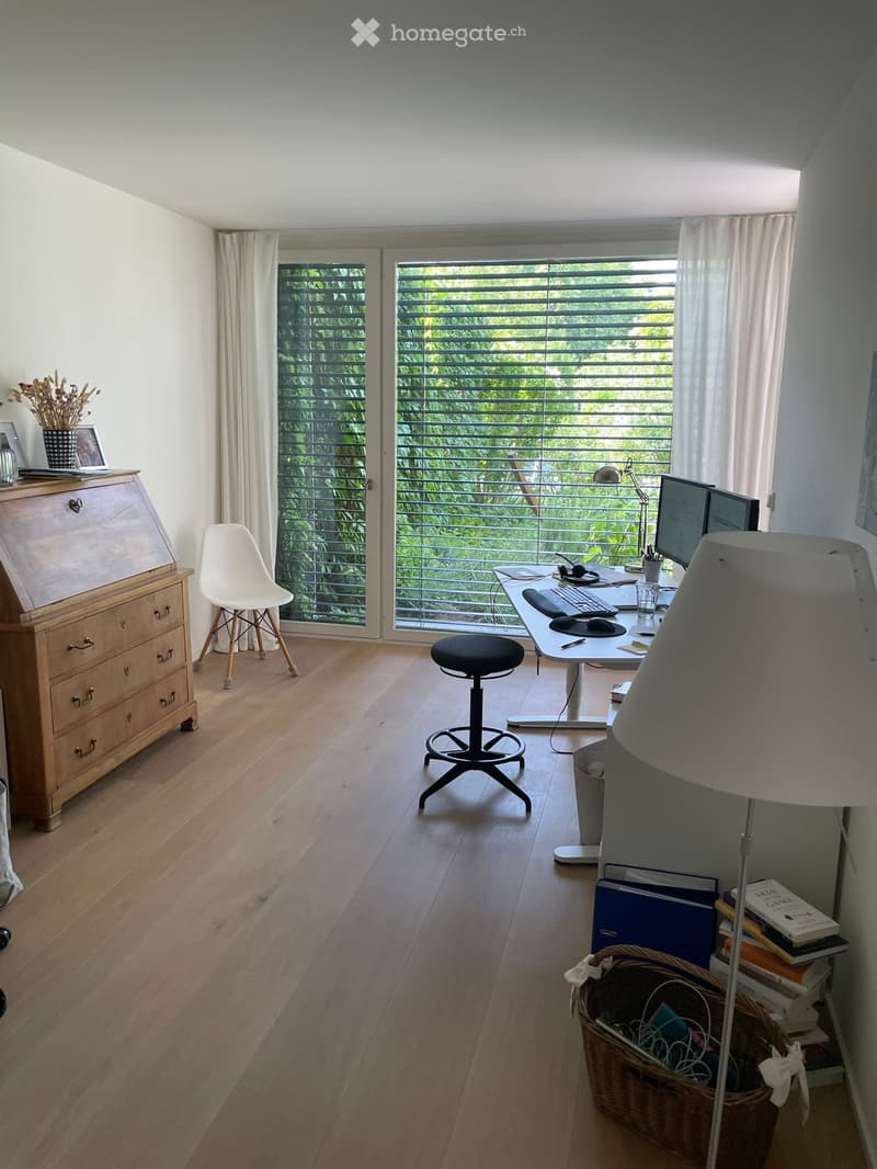 A well-lit, modern home office with wooden flooring, large windows with blinds, and a desk setup including a computer, keyboard, mouse, and lamp. There is a white plastic chair, a black stool, a wooden cabinet, and a wicker basket with items on the floor.