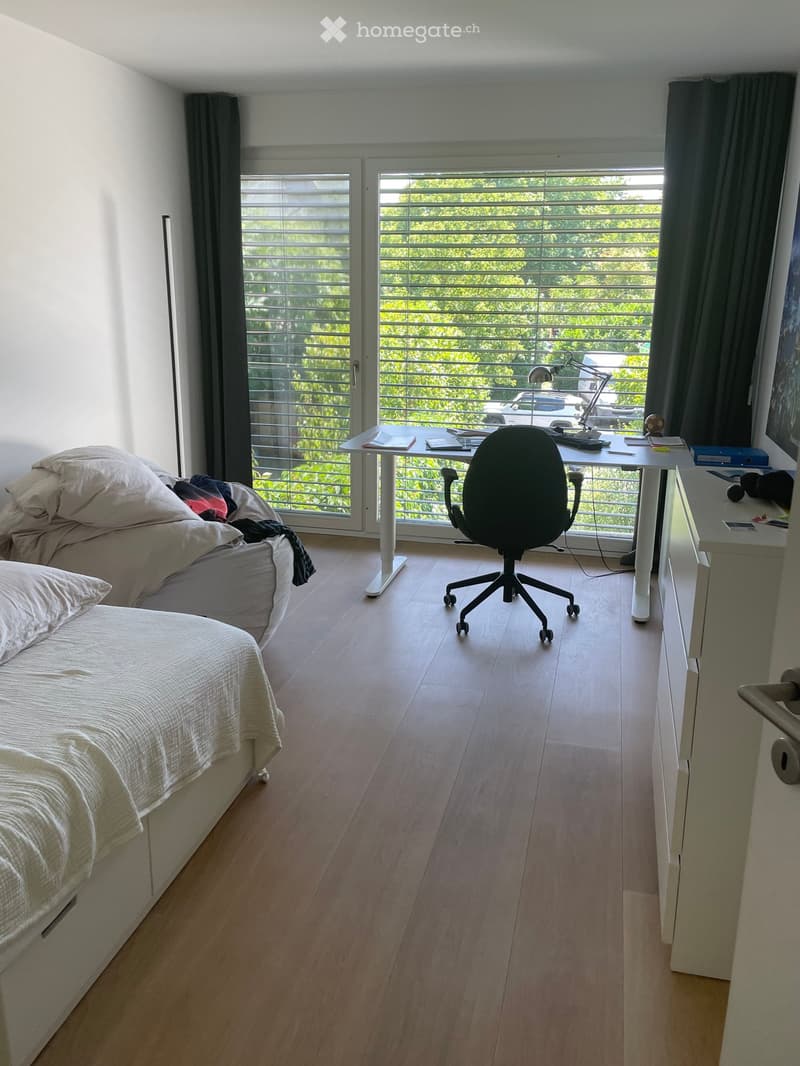 Bedroom with a bed, desk, chair, and large windows with blinds. The room features light wooden flooring and modern decor.