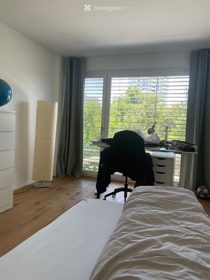 Bright bedroom with wooden flooring, large window with blinds, bed with white sheets, desk with chair, floor lamp, and globe on a cabinet.