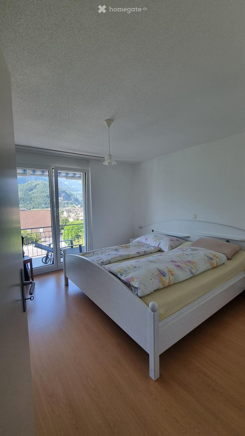 Bright bedroom with a white bed, colorful bedding, and a nightstand, featuring large windows with a scenic mountain view.