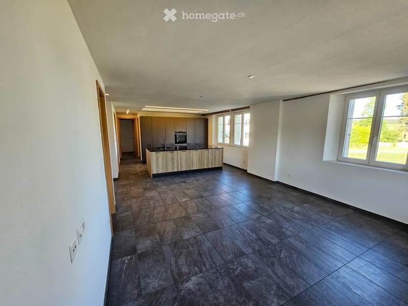 Modern kitchen with a central island, built-in oven, and sink, featuring large windows and tiled flooring.